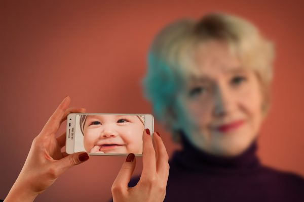 A person holds a smartphone showing a baby’s face, aligning it with the blurred face of an elderly woman in the background—a striking Fortune Cookie Friday moment, contrasting youth and age.
