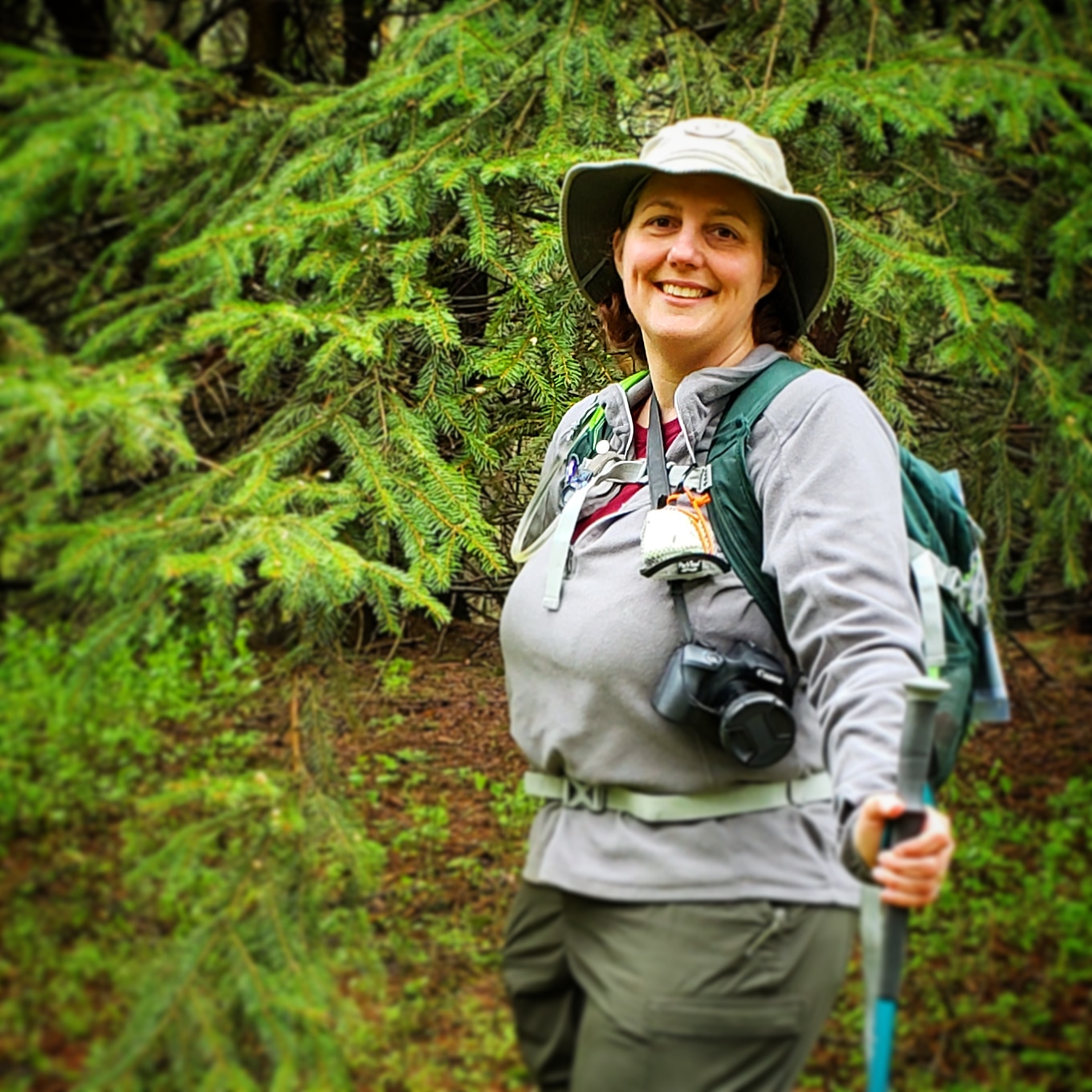 IMG_20200518_145710_839 A person wearing a hat and backpack smiles while hiking in a forest, holding a trekking pole. Pine branches surround them, and camera gear hangs from their neck.